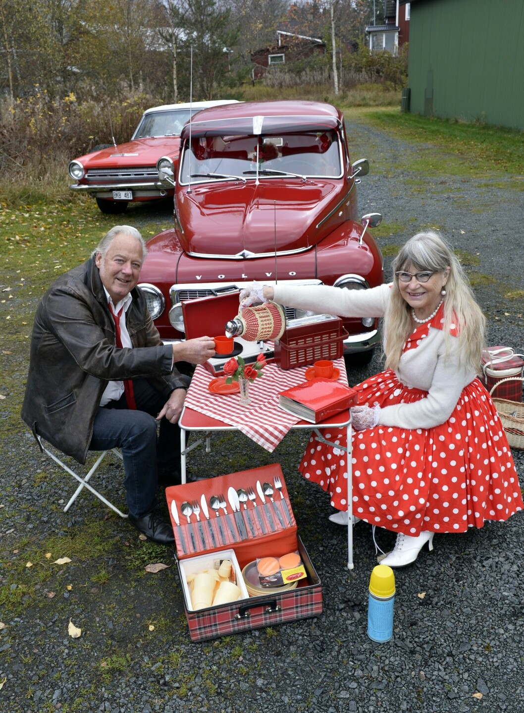 Makarna Pettersson sitter vid ett rött retrocampingbord framför sina två röda veteranbilar.
