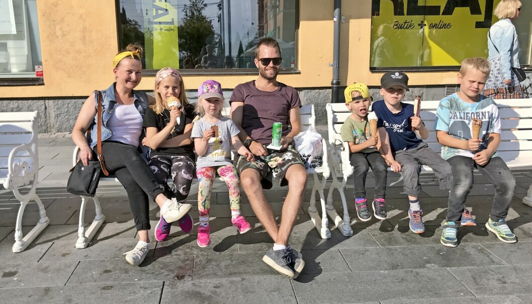 Suvi och familjen bestående av make och fem barn sitter utomhus på två vita bänkar och äter glass.