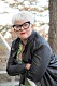 Sexologen Suzann Larsdotter lutar sig mot ett räcke utomhus. Hon bär markanta, svarta glasögonbågar, rött läppstift, svart skinnjacka och en mönstrad klänning i grönt, svart och vitt.