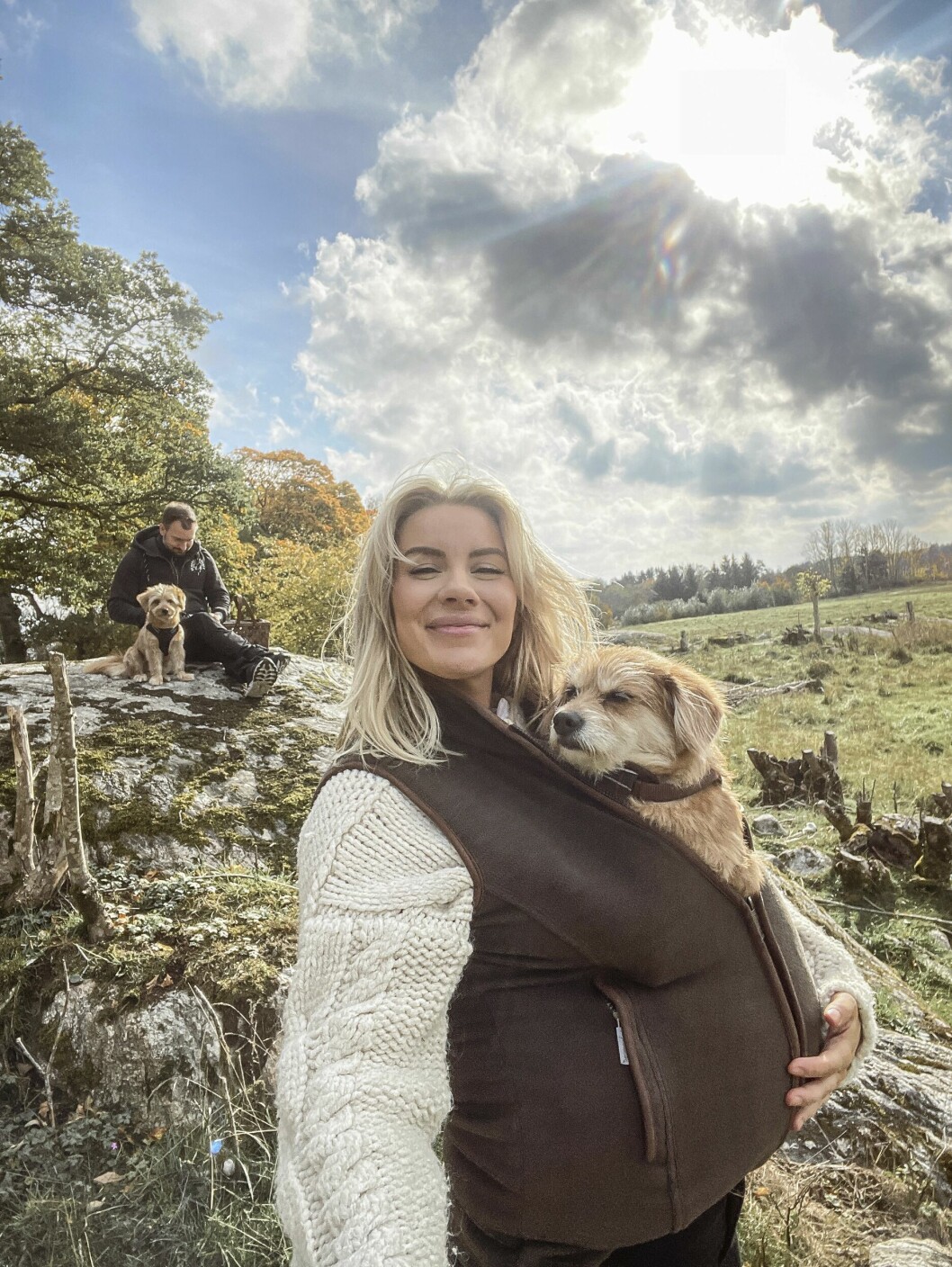 Therése Lindgren ute i naturen med Anders Vesterlund och deras hundar. Han sitter med den ena hunden på en klippa i bakgrunden, hon bär den andra innanför västen.
