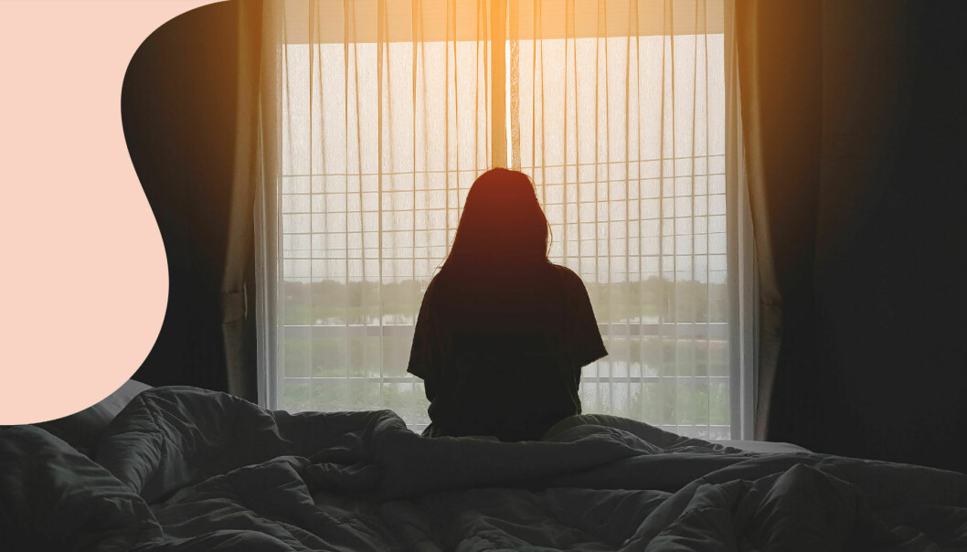 En kvinna sitter i motljus på en säng med ryggen mot kameran och tittar ut genom fönstret.