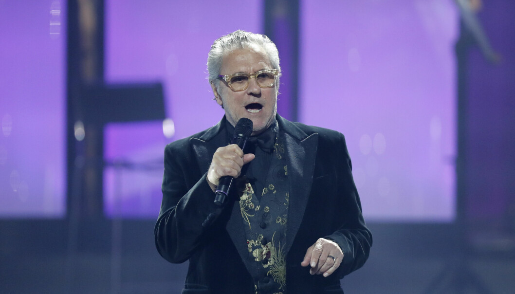 Tommy Körberg står på en scen och sjunger i en mikrofon. Han är klädd i en svart kavaj och en väst i svart och guld. Han har glasögon.