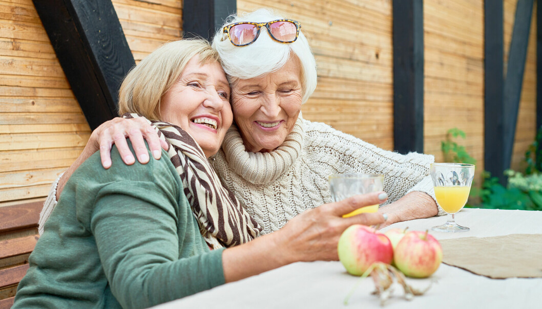 Två äldre, glada, kvinnor sitter utomhus och håller om varandra samtidigt som de skrattar.