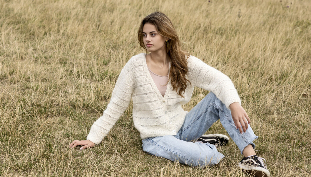 Ung kvinna i jeans och vit kofta sitter i gräset.